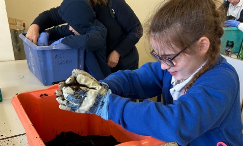Close-up of a girl wearing gardening gloves picking up soil from a big red tub
